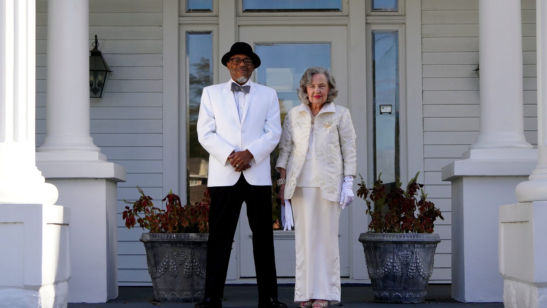 The River King in a white dinner jacket and the Moss Queen in white standing between the columns of a porch in Moss Point